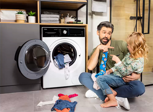 Mężczyzna siedzący z dzieckiem na kolanach robiący pranie z suszarką Amica AD3C83iLEB