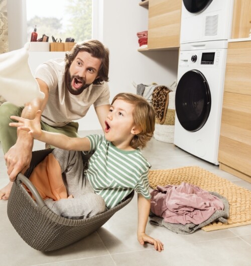 Mężczyzna bawiący się z dzieckiem siedzącym w koszu na pranie; w tle urządzenie marki Amica