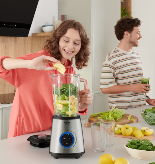 Kobieta w kuchni wrzuca owoce i szpinak do blendera Amica, mężczyzna w tle trzyma koktajl
