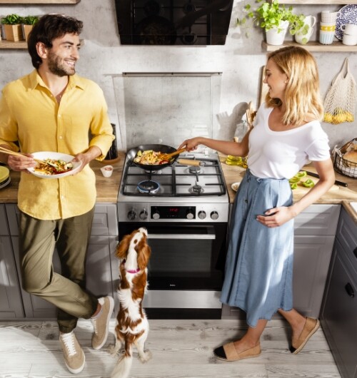 Kobieta gotująca potrawę na płycie gazowej marki Amica; obok mężczyzna i pies